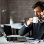 Man talking on phone, drinking coffee, looking at laptop screen