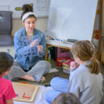 A Montessori teacher sits on the floor, guiding young students through a lesson.